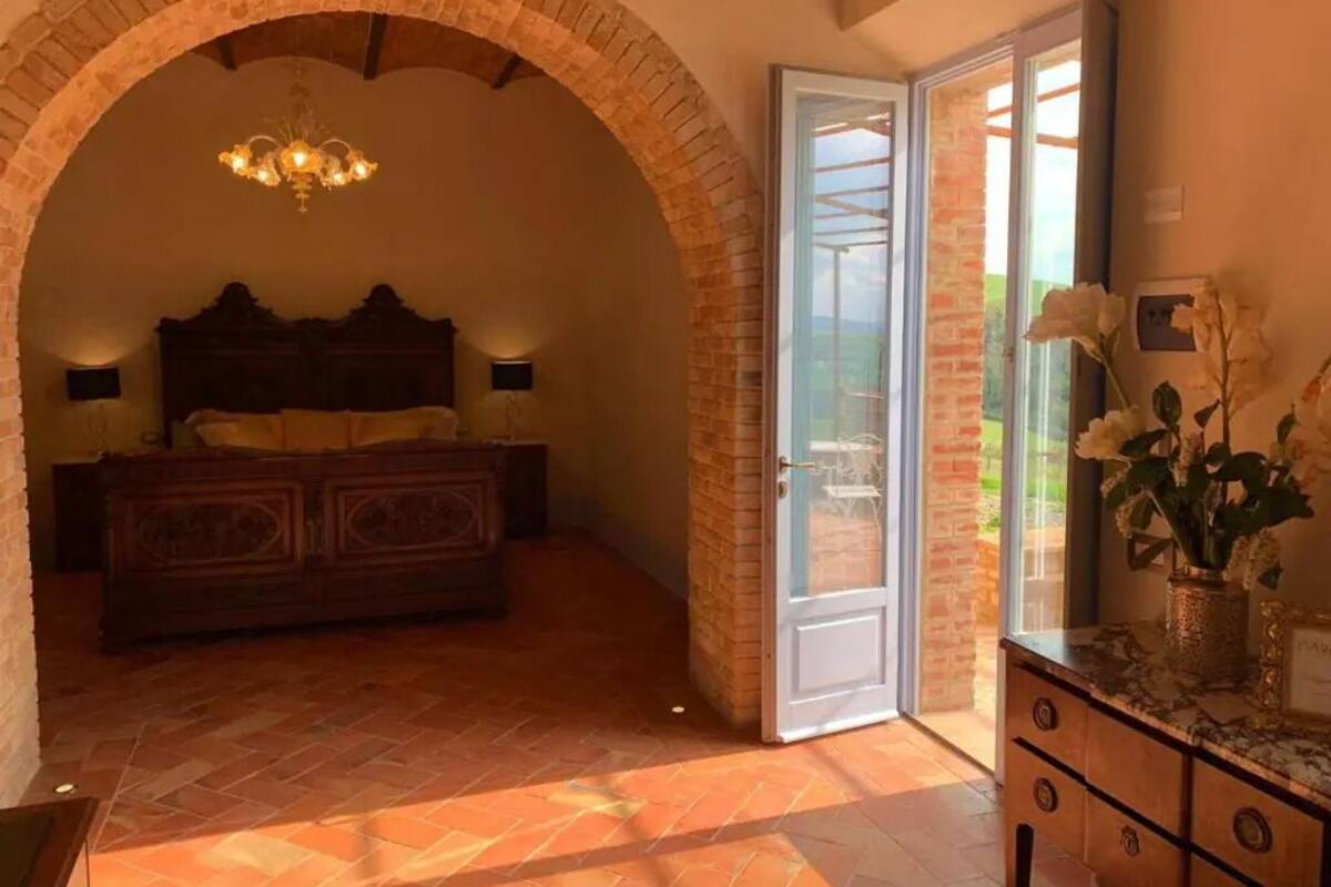 A bedroom with a brick archway, a wooden bed, a dresser, and a sliding glass door leading to a balcony.
