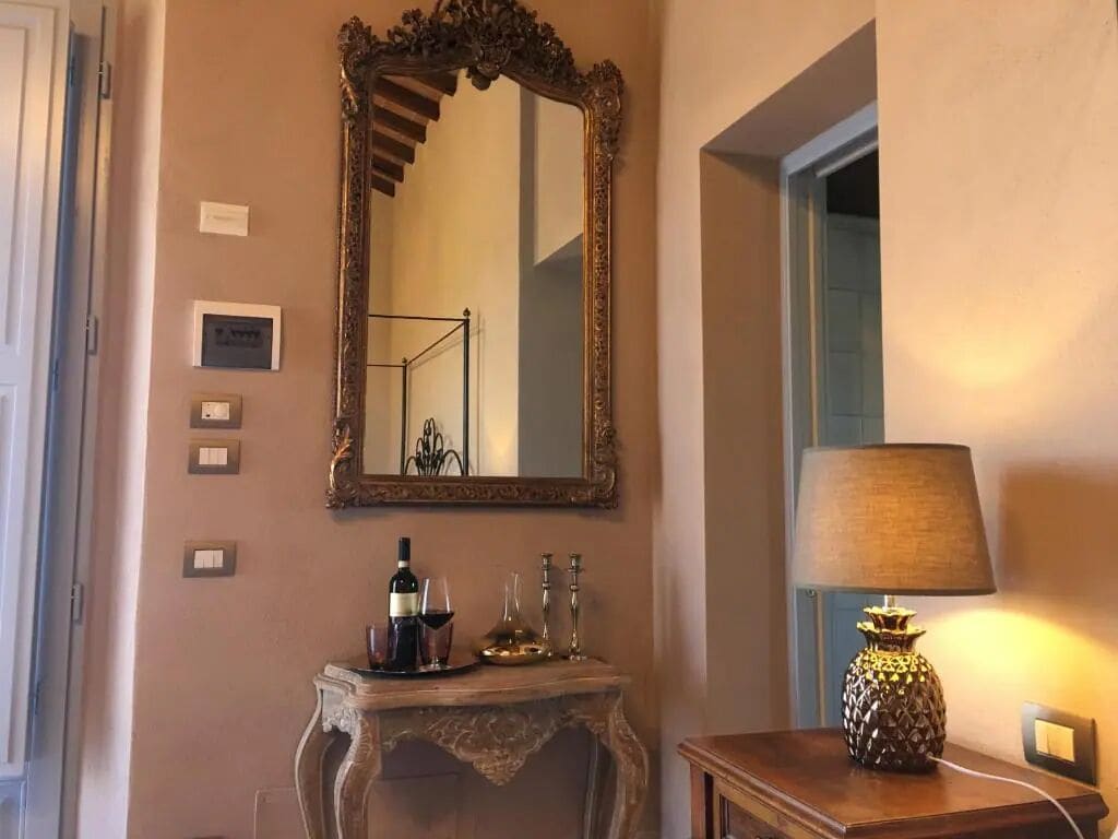 Une pièce avec un grand miroir décoratif, une console en bois sur laquelle sont posés une bouteille et des verres, et une lampe sur une petite table d’appoint.