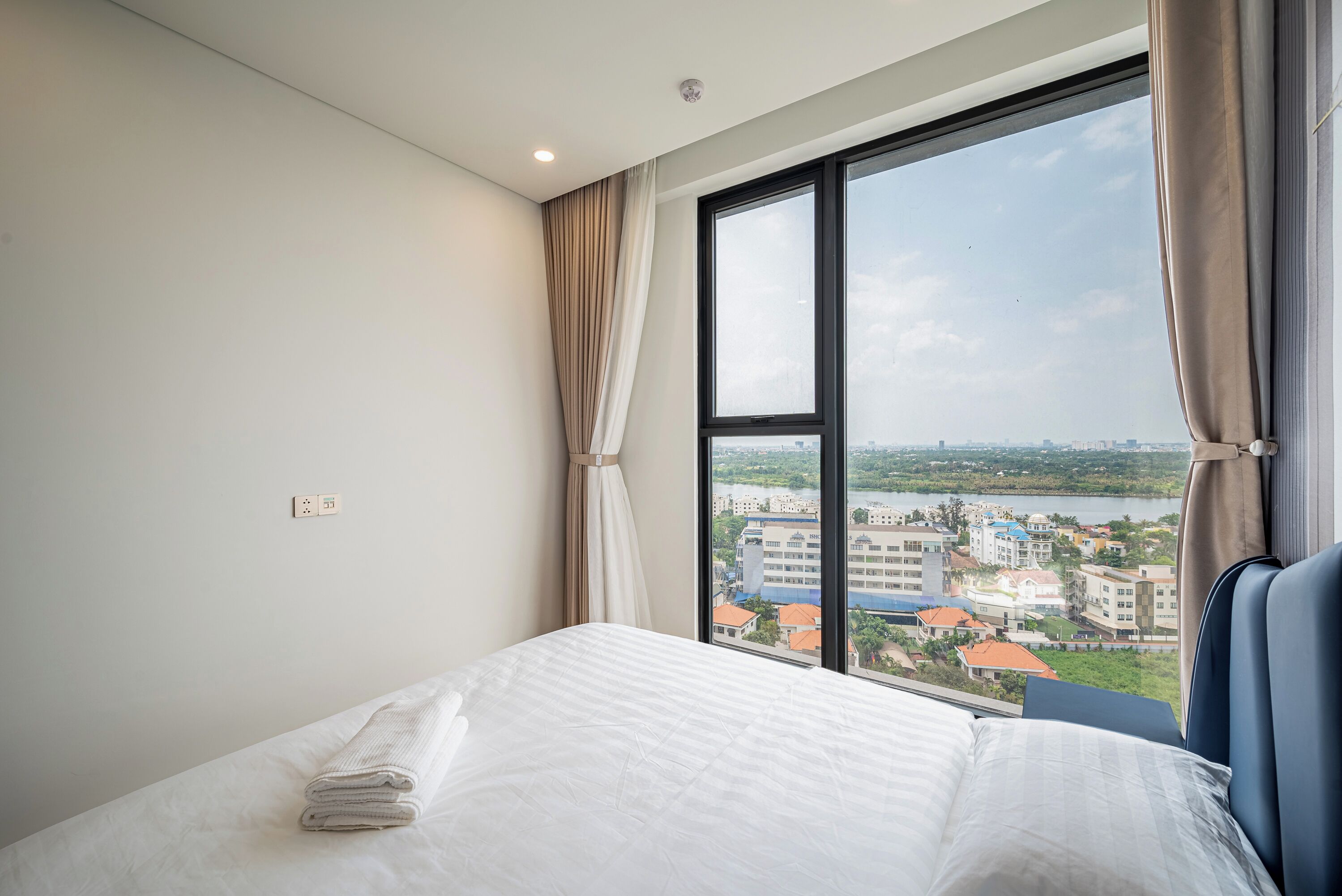 A hotel room with a large window, a neatly made bed, and a view of a cityscape and water.