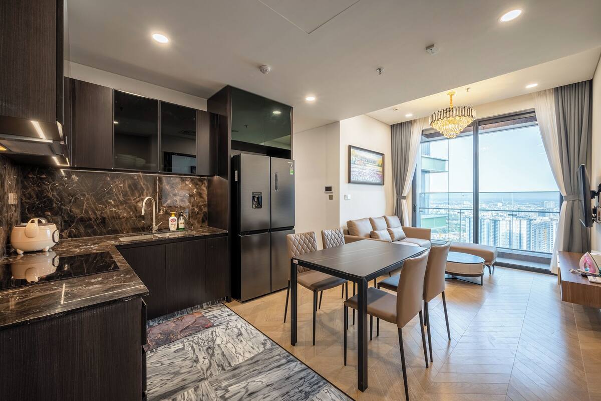 Eine moderne Küche mit dunklen Schränken, einer Marmorarbeitsplatte und einem eingebauten Kühlschrank. Es gibt einen Essbereich mit Tisch und Stühlen sowie ein großes Fenster mit Blick auf die Stadt.