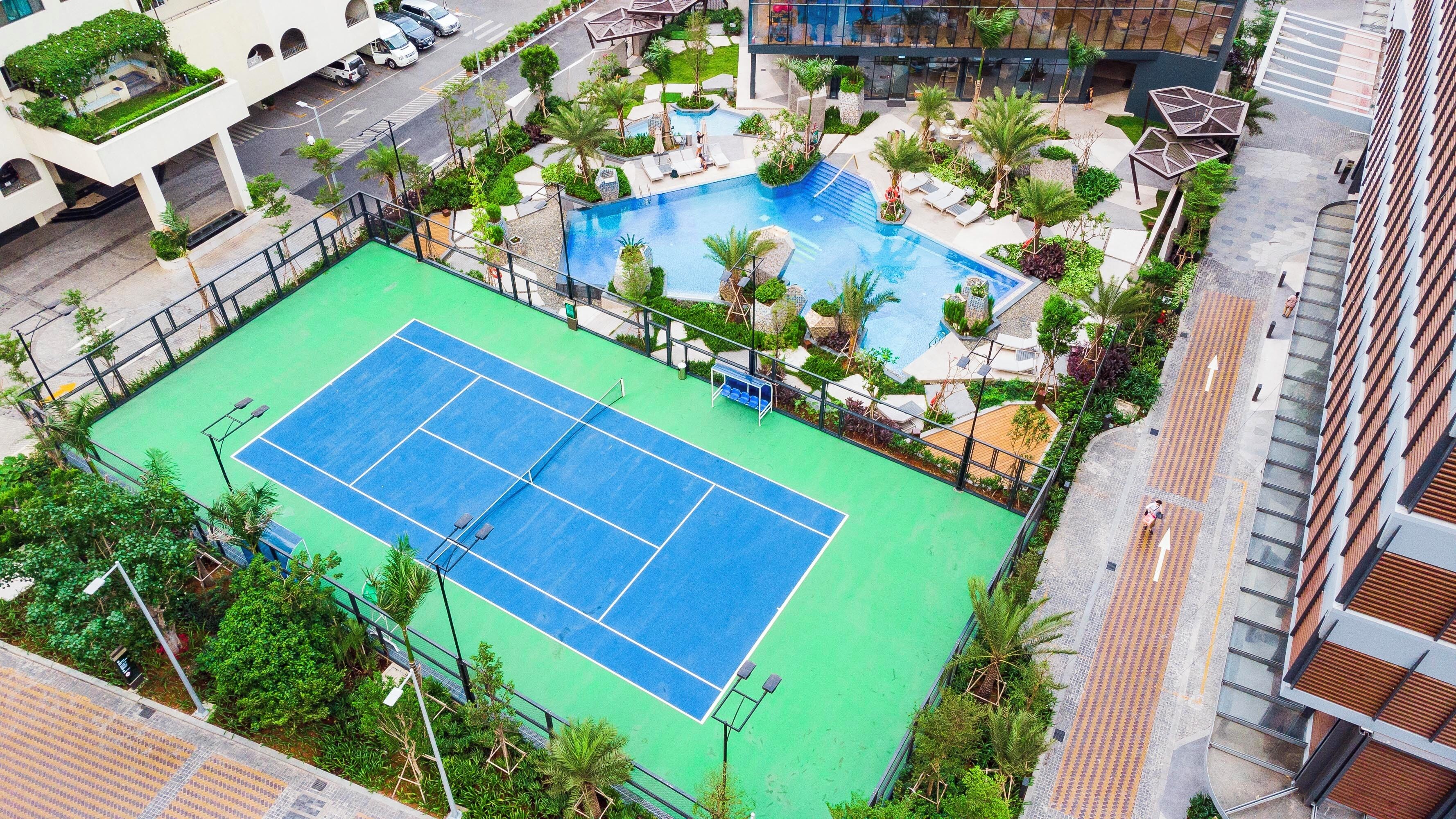 An aerial view of a tennis court surrounded by greenery, with a pool and lounge area nearby.