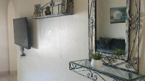 A living room with a mounted TV, a decorative mirror, and a glass shelf with plants. - EL MANZEL 102 (HOUMT SOUK)