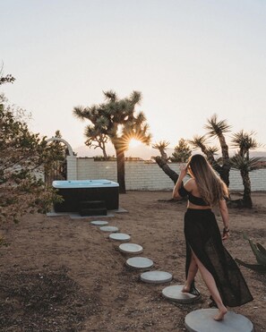 Une femme marchant sur des pierres plates qui lui servent de marchepied, en se dirigeant vers un bain à remous dans un jardin désertique, avec des cactus et un coucher de soleil en arrière-plan.