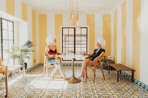 Une pièce avec un sol carrelé, une table ronde et des chaises. Il y a une fenêtre avec des rayures verticales sur le mur.