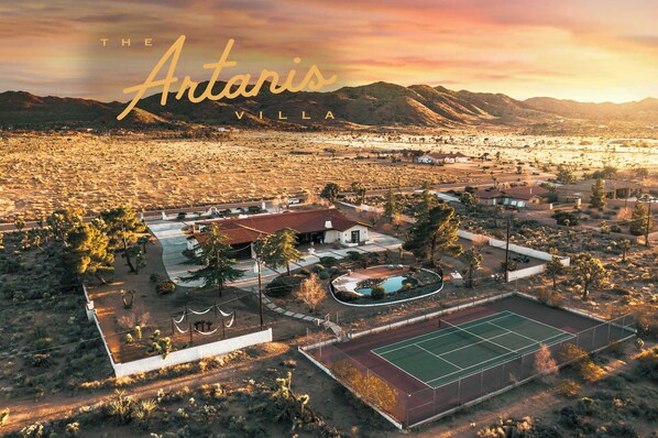 An aerial view of The Antigua Villa, featuring a tennis court, swimming pool, and surrounding landscape with mountains in the background.