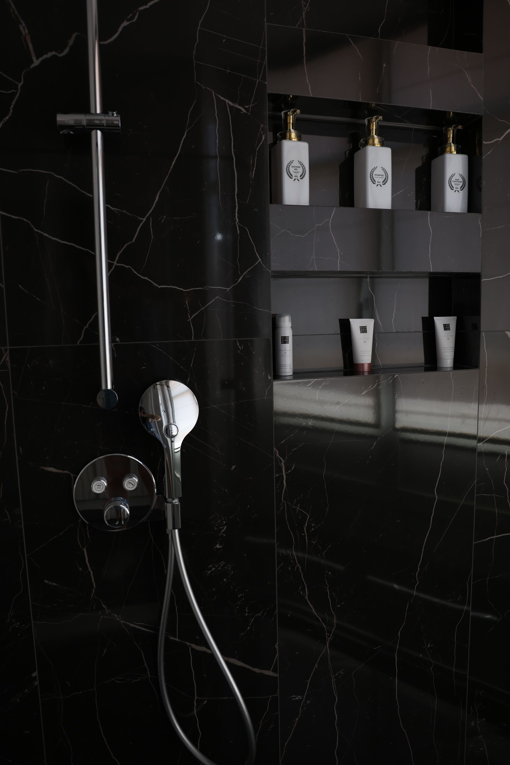 Une salle de bain moderne avec un mur en marbre noir, une pomme de douche chromée et des étagères contenant des articles de toilette.