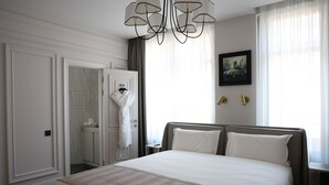 Habitación de hotel con una cama grande, un candelabro, un cuadro en la pared y un baño visible a través de una puerta abierta.