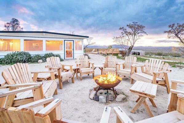 A beachside outdoor seating area with wooden Adirondack chairs and a central fire pit.