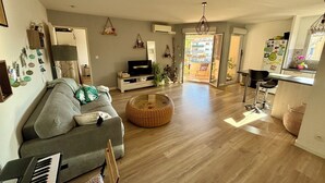 Un salon avec un canapé gris, un pouf en osier, un piano et un téléviseur.
