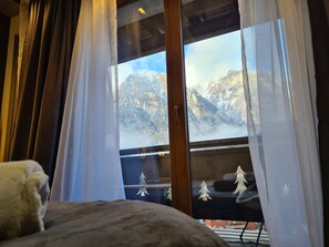 Een kamer met een groot raam, gordijnen, een bed en uitzicht op besneeuwde bergen.