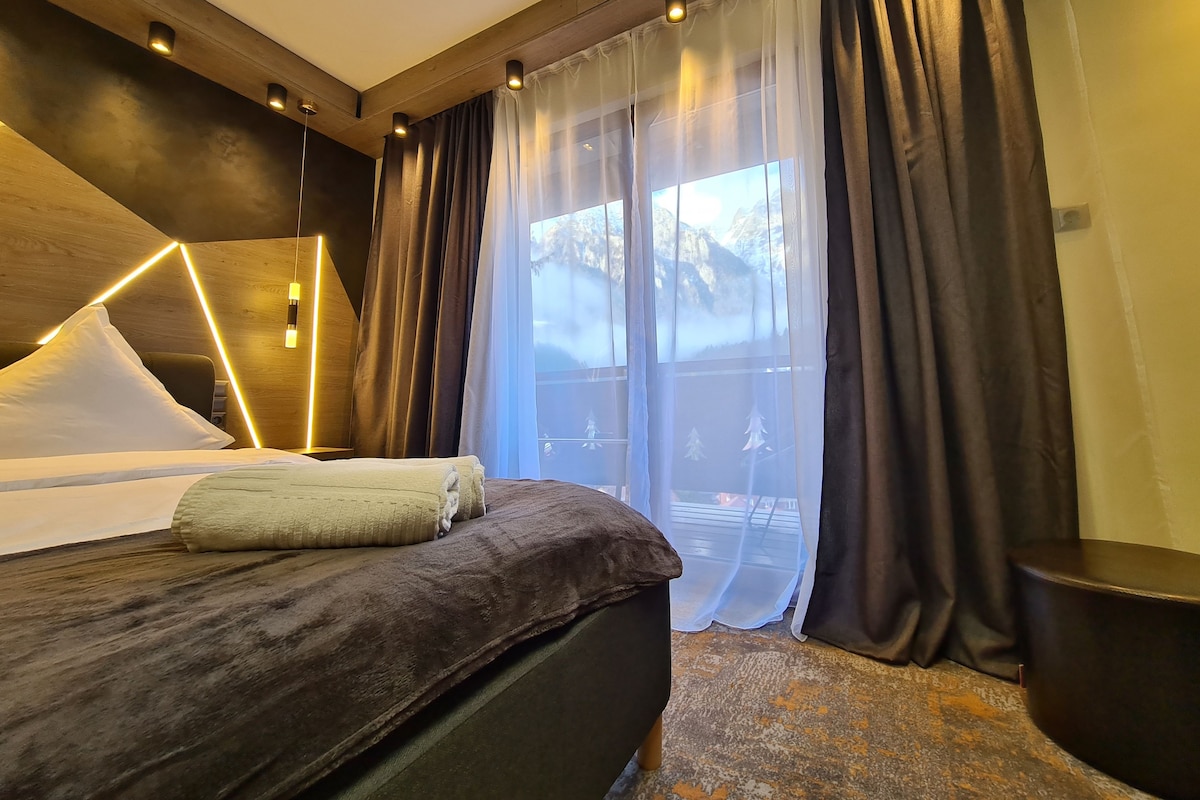 A modern hotel room with a large bed, a wooden headboard, and a view of a mountainous landscape through sheer curtains.