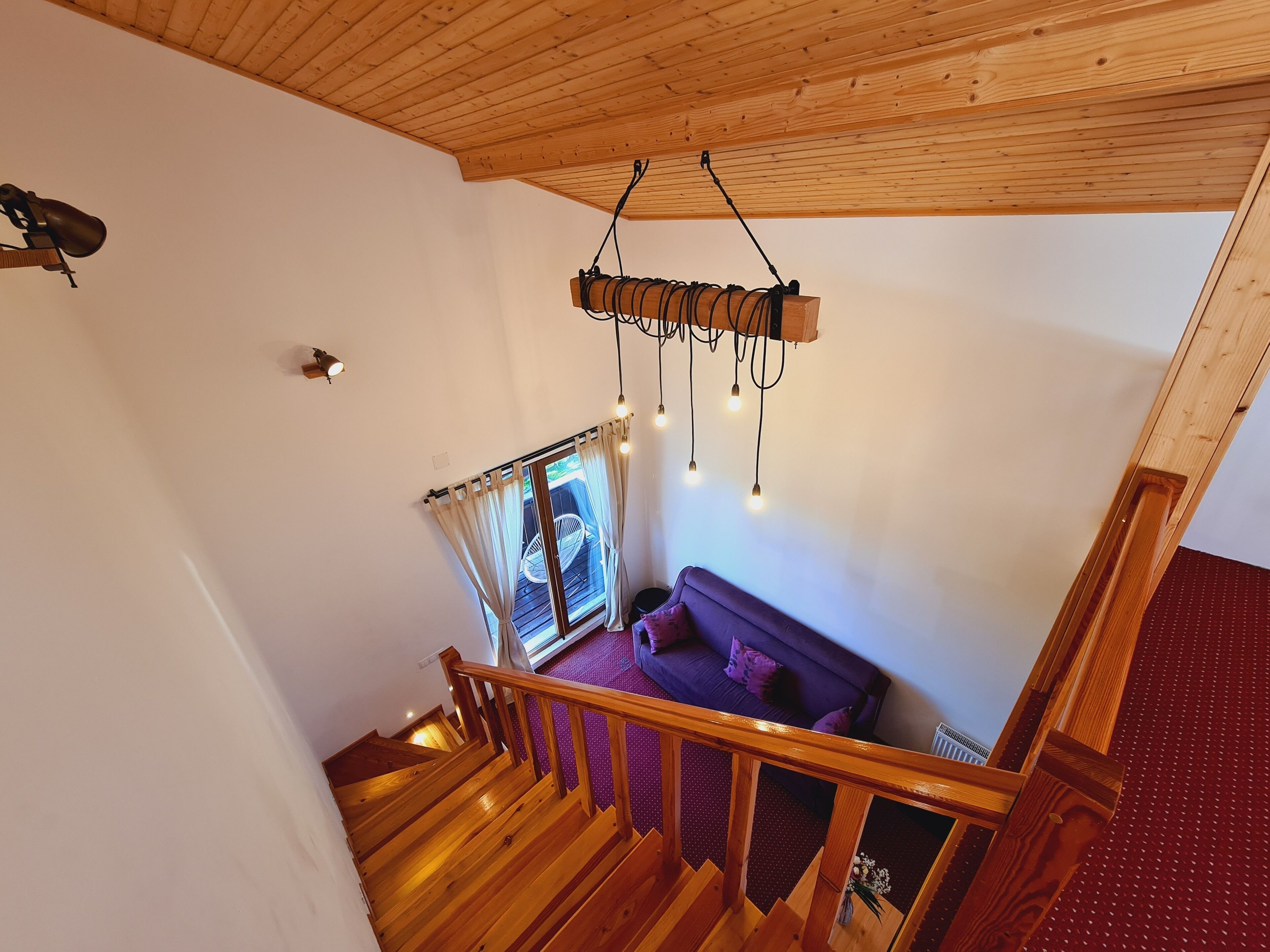 Un escalier en bois mène à une pièce où se trouvent un canapé violet, une fenêtre avec des rideaux et un luminaire suspendu.