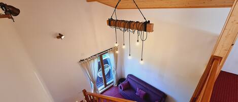 Un escalier en bois mène à une pièce où se trouvent un canapé violet, une fenêtre avec des rideaux et un luminaire suspendu.