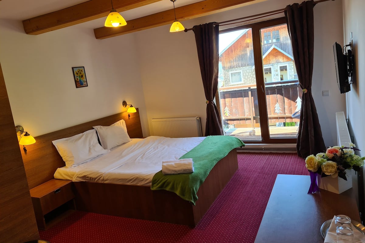 A hotel room with a wooden bed, a red carpet, a window with curtains, and a view of a building outside.