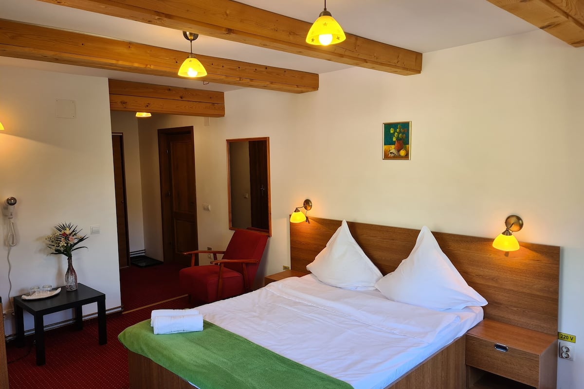 A hotel room with a bed, bedside lamps, a wooden headboard, a small table with a vase, a red chair, and a painting on the wall.