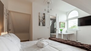 A hotel room with a bed, a brown blanket, a white towel, a chandelier, a painting, a window, and a plant.
