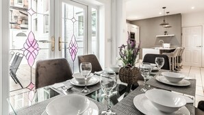 A dining area with a glass table, white plates, and clear glasses. There are chairs around the table, and a kitchen is visible in the background.