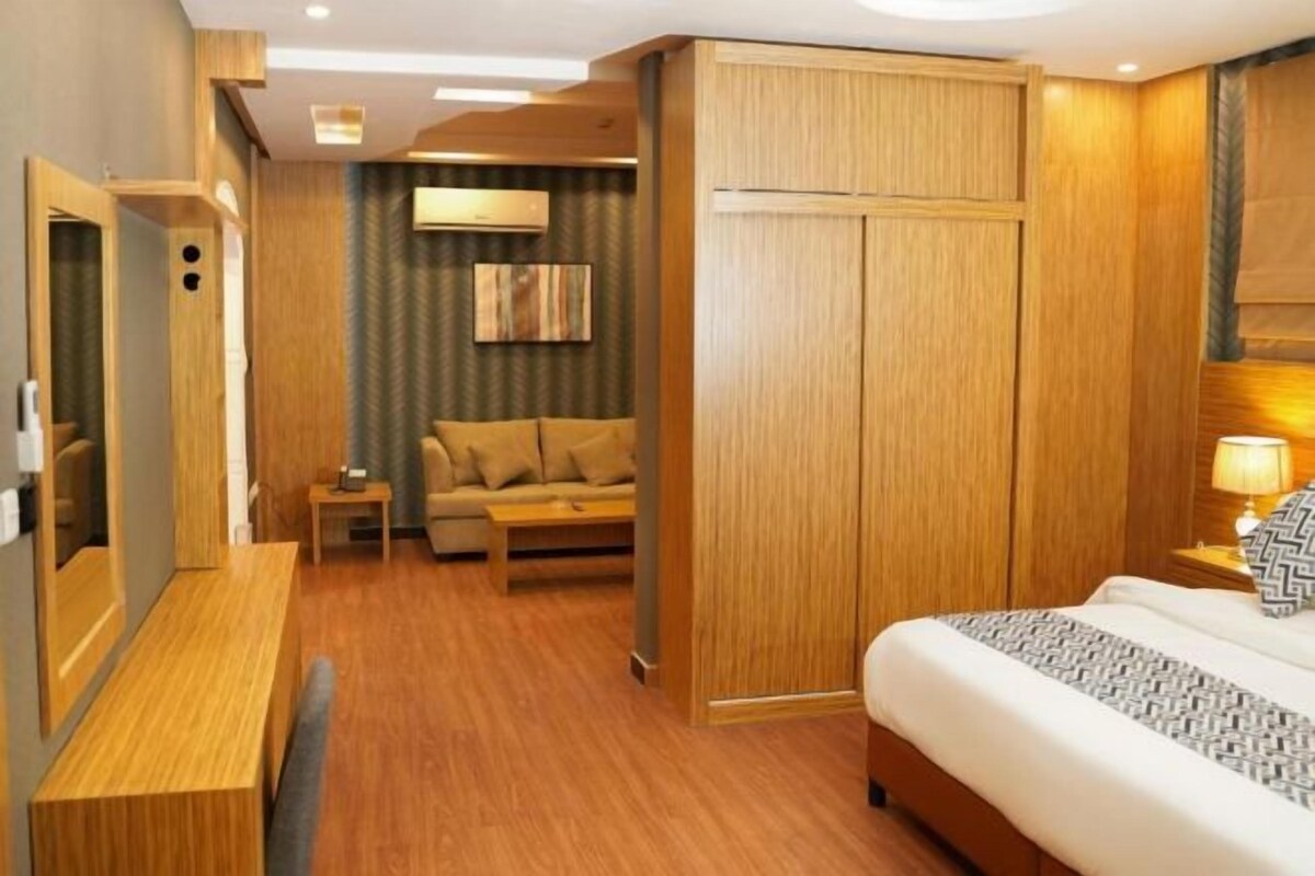 Habitación de hotel con piso de madera, cama con colcha estampada, mesita de noche con lámpara, sofá y televisor instalado en la pared.