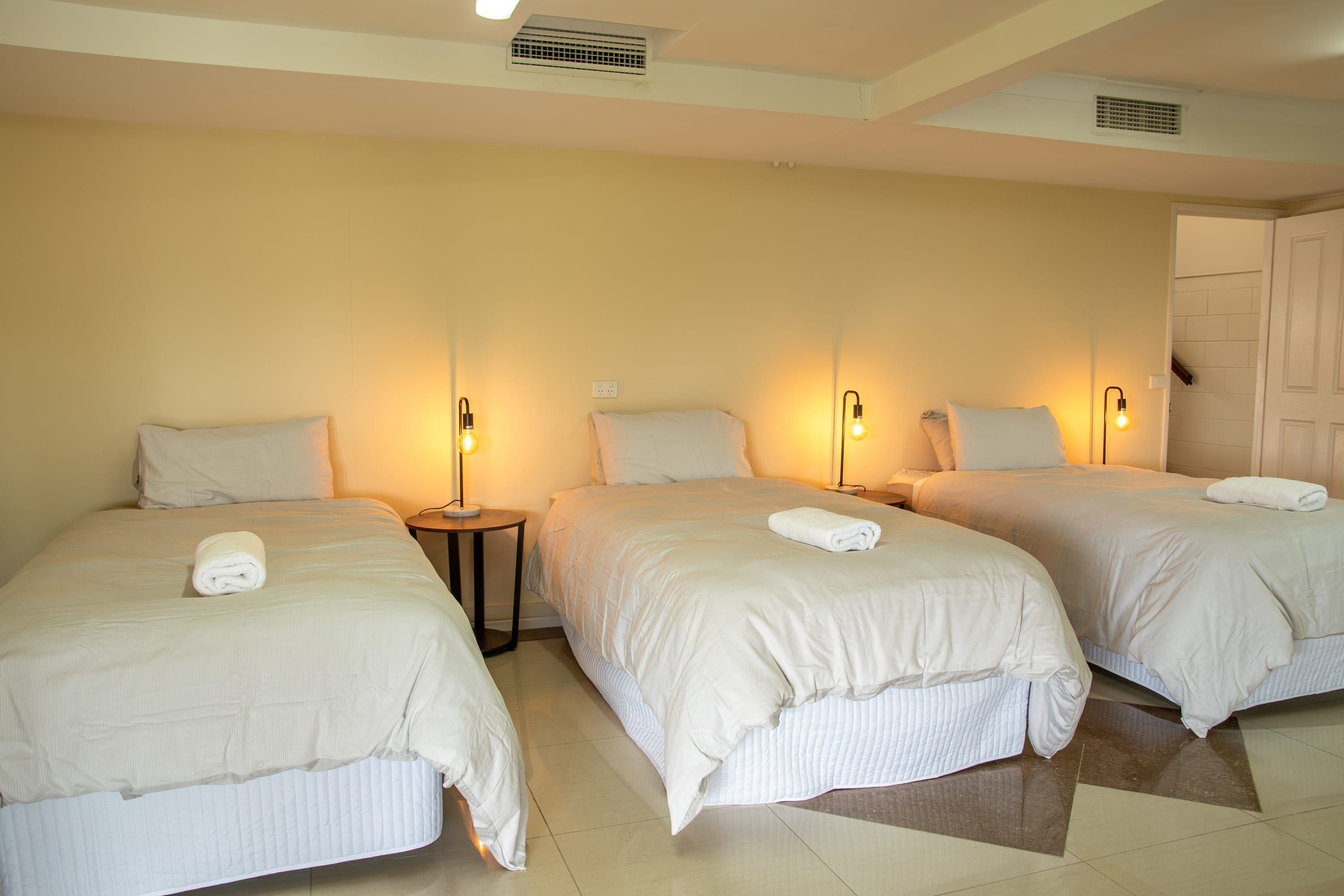 A hotel room with three beds, each with a white duvet and a rolled towel on top. There are bedside lamps on each bed, and a door to the right.