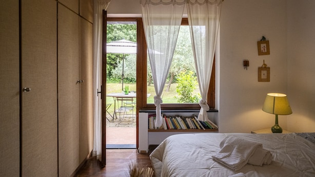 A bedroom with a bed, a bedside table with a lamp, a window with sheer curtains, and a view of an outdoor patio area.