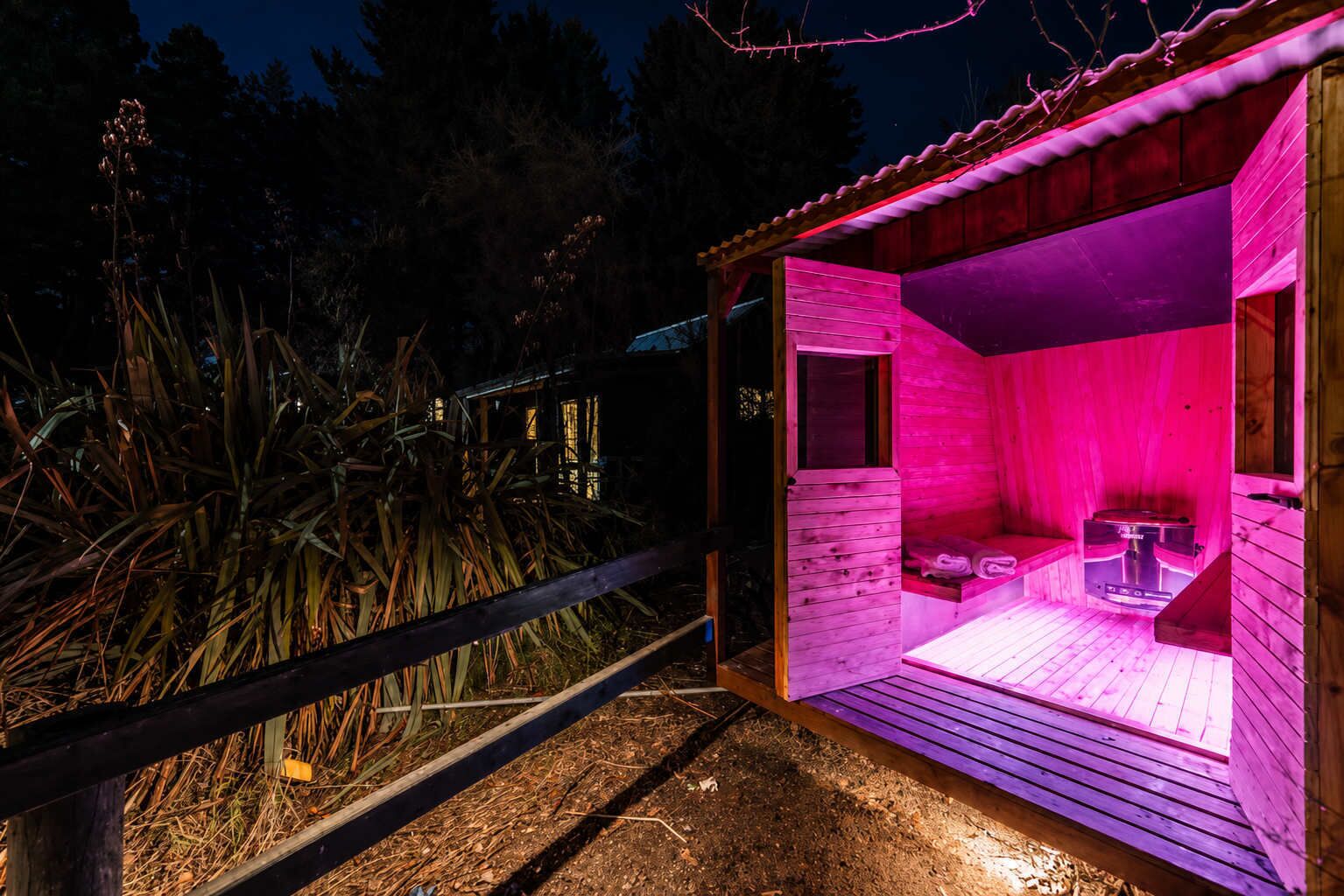 A wooden sauna with a bench and a table, surrounded by a wooden fence and vegetation.
