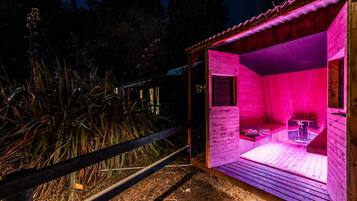 A wooden sauna with a bench and a table, surrounded by a wooden fence and vegetation.