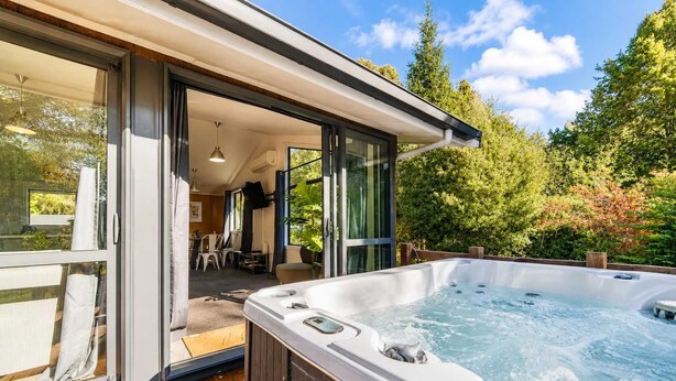 Ein moderner Außenbereich mit Whirlpool, einer Holzterrasse, einer Glastür, die zu einer Terrasse führt, und Blick auf einen üppigen Garten.