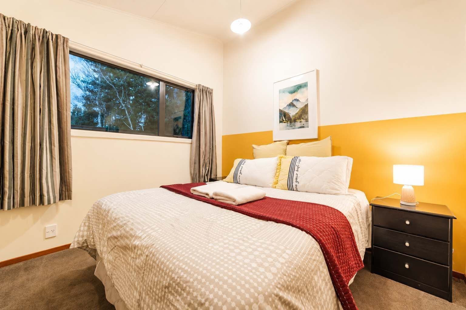 A hotel room with a bed, a nightstand, a lamp, a window with curtains, and a painting on the wall.