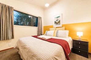 Una habitación de hotel con cama, mesita de noche, lámpara, ventana con cortinas y un cuadro en la pared.