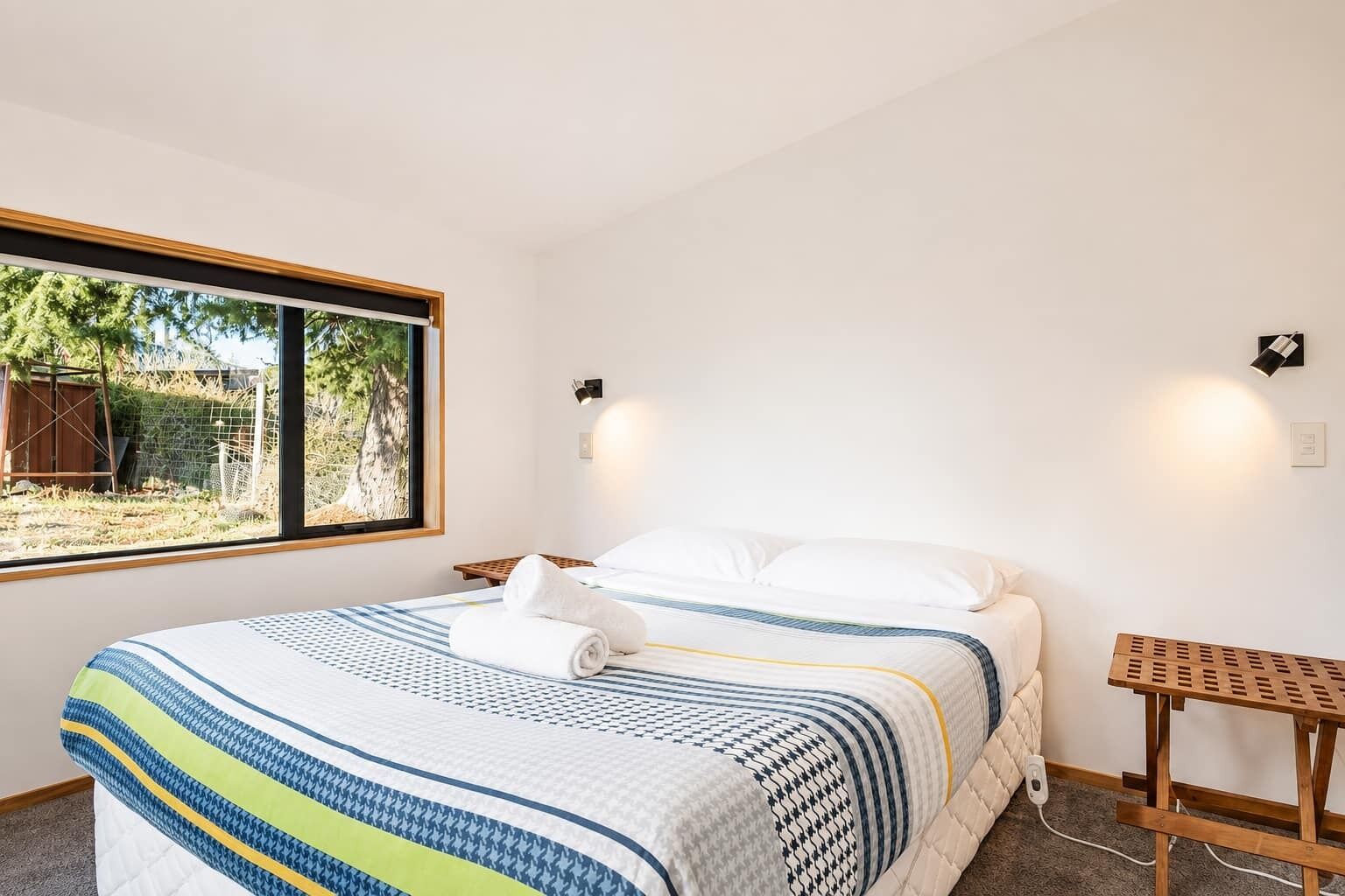 A neatly made bed with a striped bedspread, a bedside table, and a window with a view of greenery outside.