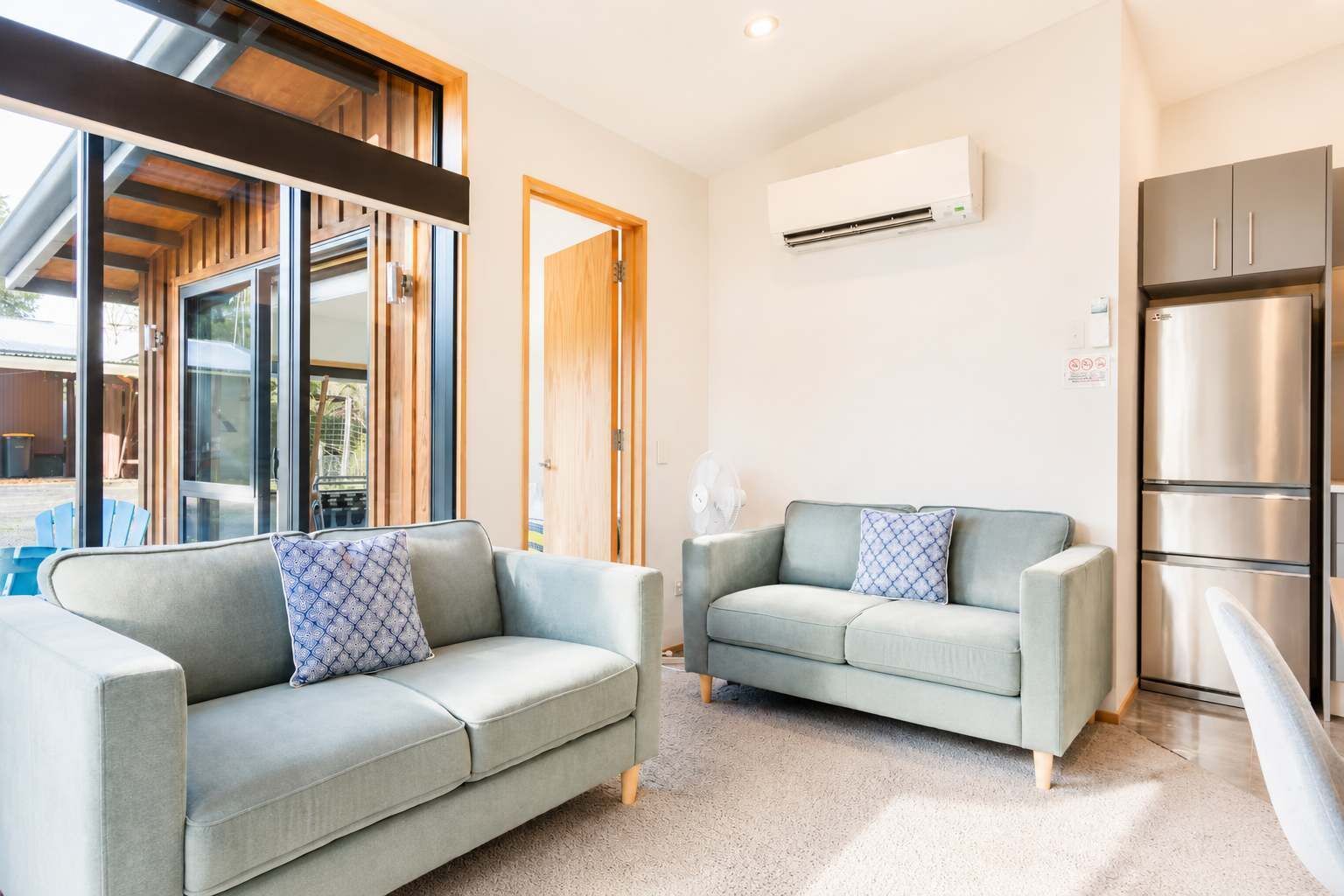 A modern living room with two grey sofas, a large window, an air conditioning unit, and a built-in stainless steel refrigerator.