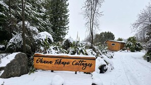 A wooden sign for Ohana Tekapo Cottages with a snow-covered landscape and trees in the background.