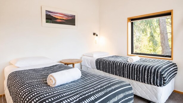 A hotel room with two beds, a small table, a wall-mounted light, a framed picture, and a window with a view of trees.