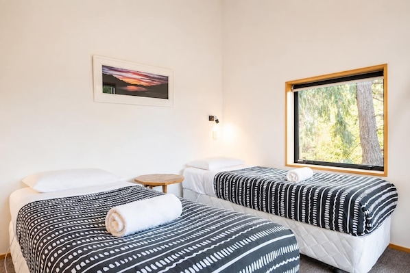 Habitación de hotel con dos camas, una mesita, una lámpara de pared, un cuadro enmarcado y una ventana con vista a árboles.