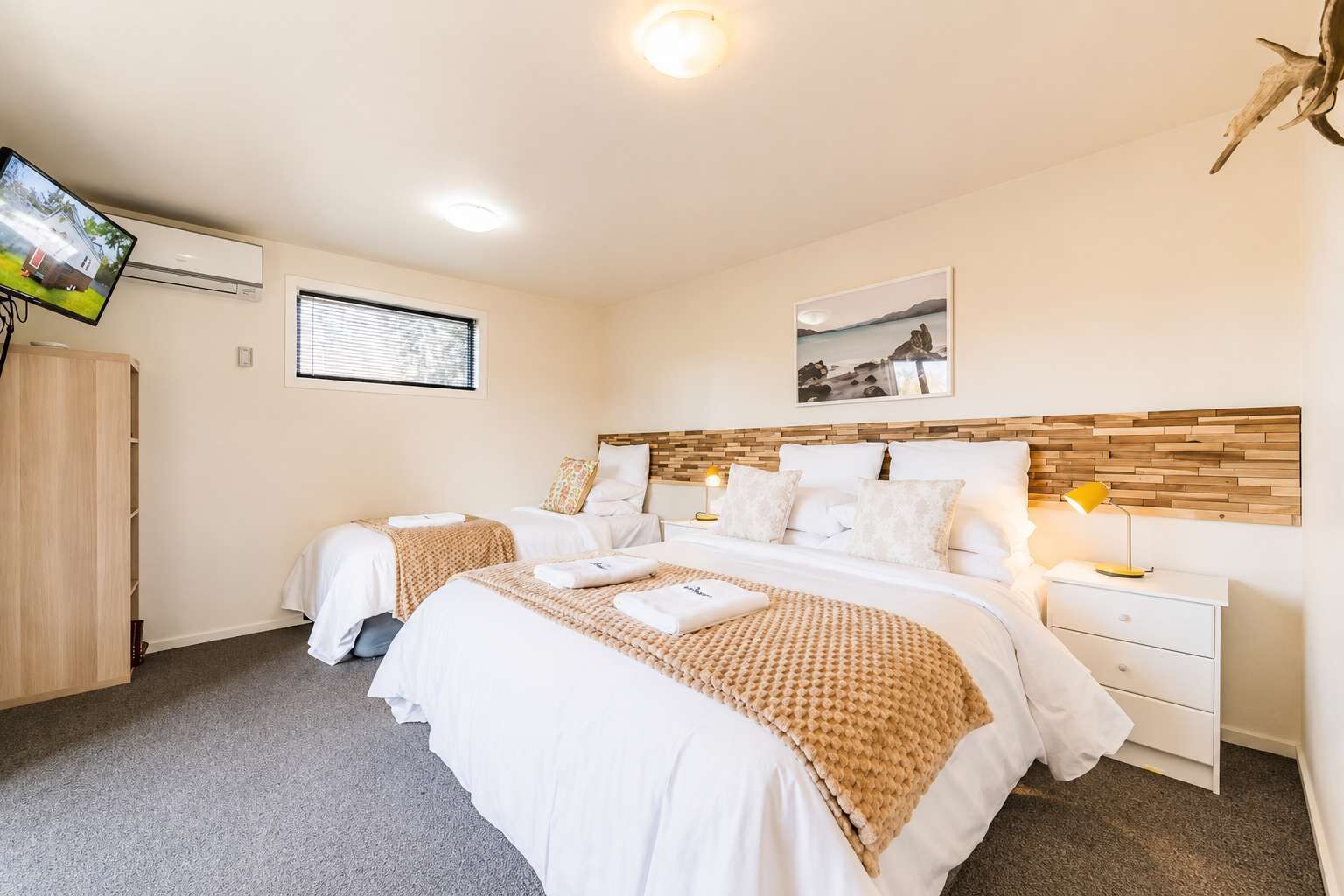 A hotel room with two beds, a TV, a wooden headboard, a bedside table with a lamp, and a picture on the wall.