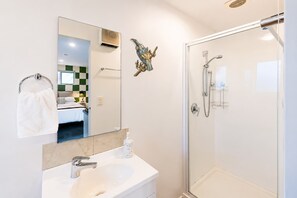 A modern bathroom with a glass shower enclosure, a white sink, and a mirror reflecting a room with a bed and a green and white checkered wall.