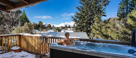 A person in a hot tub on a wooden deck with a snowy mountain backdrop.