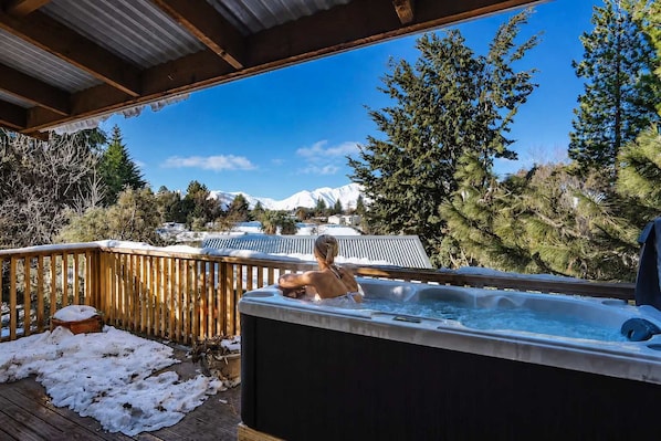 A person in a hot tub on a wooden deck with a snowy mountain backdrop.