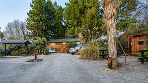 A gravel driveway leading to a wooden cabin with a metal roof, surrounded by tall trees and a small wooden structure. - Milkyway Lodge | Hot Tub + Scenic Views (Lake Tekapo)