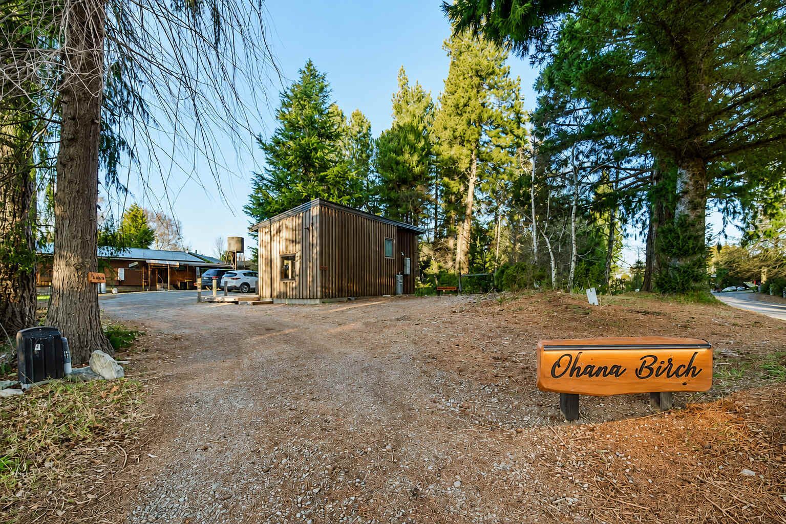Eine rustikale Holzhütte mit Schotterweg, umgeben von hohen Bäumen und blauem Himmel.