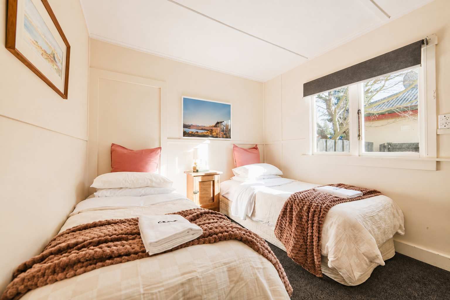A hotel room with two beds, a nightstand, a window with a view, and a painting on the wall.