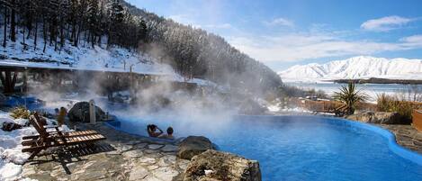A hot spring with a stone pool, wooden lounge chairs, and a forested mountain backdrop.