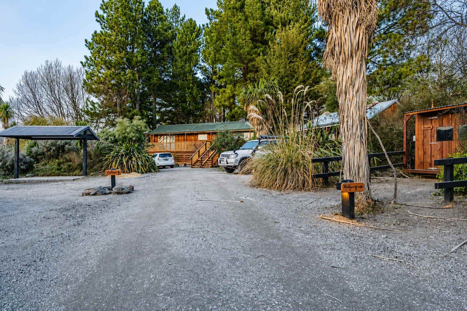 A gravel driveway leading to a wooden cabin with a metal roof, surrounded by tall trees and a small wooden structure.
