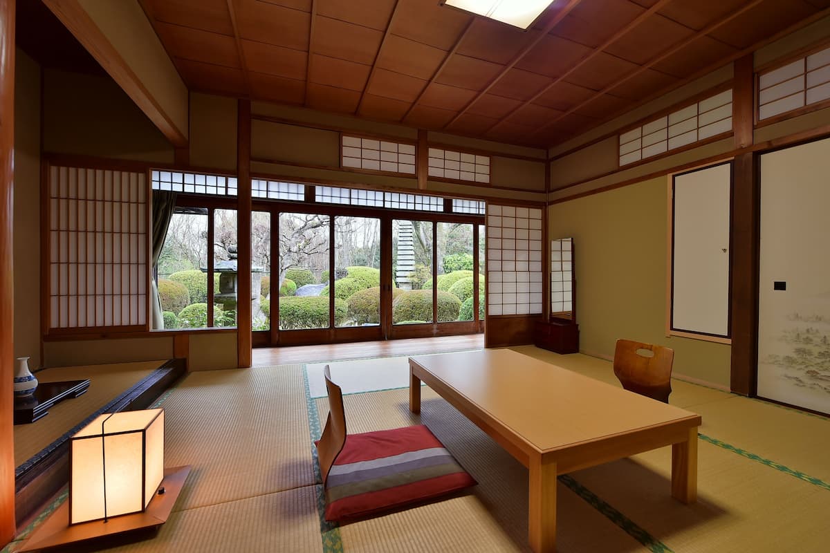 A traditional Japanese room with tatami flooring, a low table, a cushion, and sliding doors with a view of a garden.
