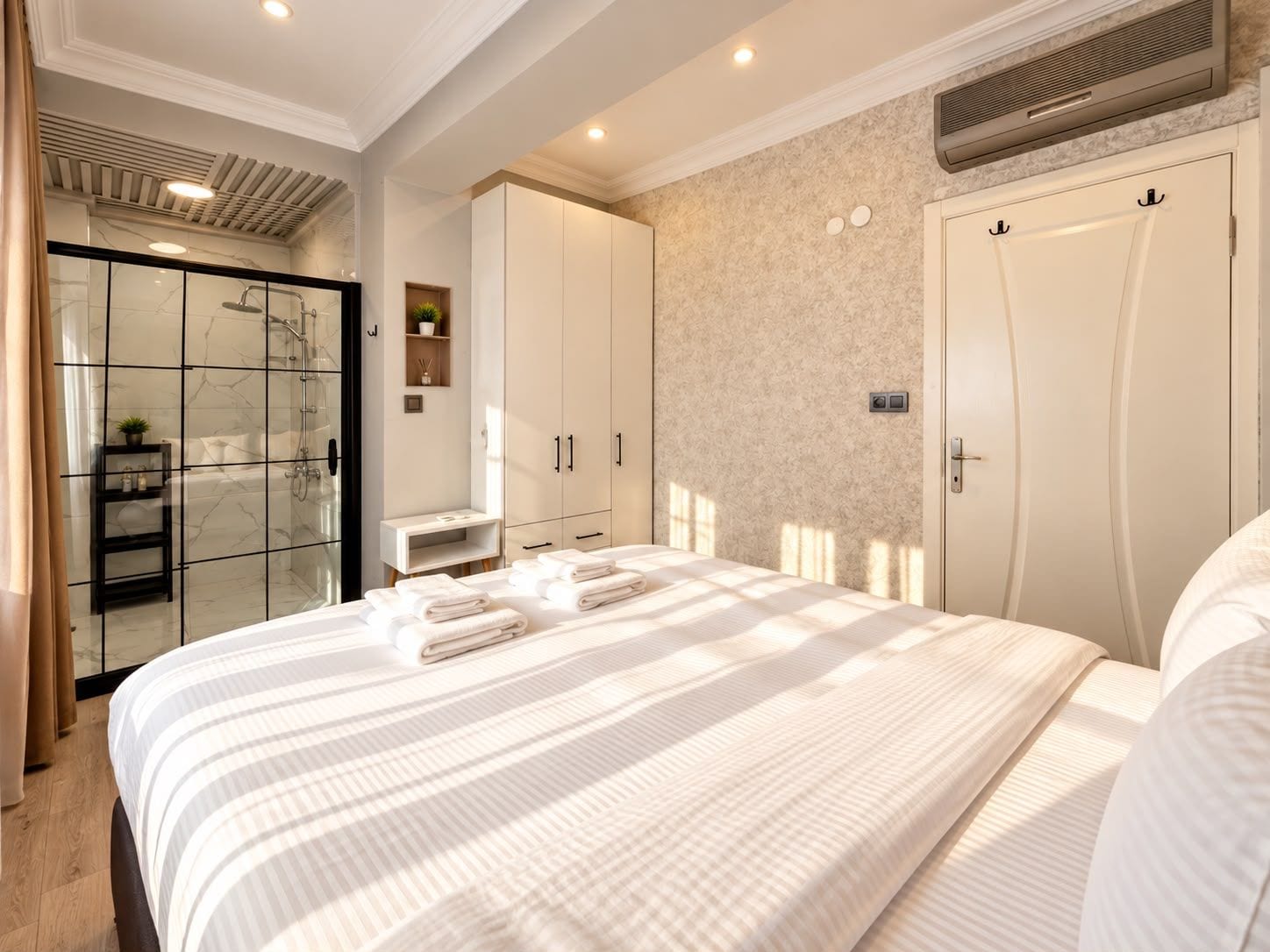 A hotel room with a large bed, a walk-in closet, a bathroom with a glass shower enclosure, and a wall-mounted air conditioning unit.
