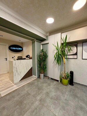 Un área de recepción con mostrador de mármol, un letrero que dice 'CASA FLORYA' y varias plantas en macetas.