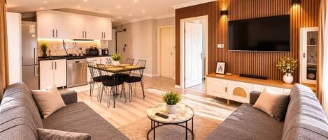 A modern living room with a grey sofa set, a dining area with a table and chairs, a flat-screen TV mounted on a wooden wall, and a kitchen in the background.