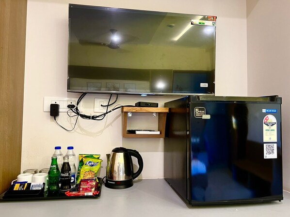 A kitchenette with a flat-screen TV mounted above a black refrigerator, a coffee maker, and a shelf with bottled water, soda, and snacks.