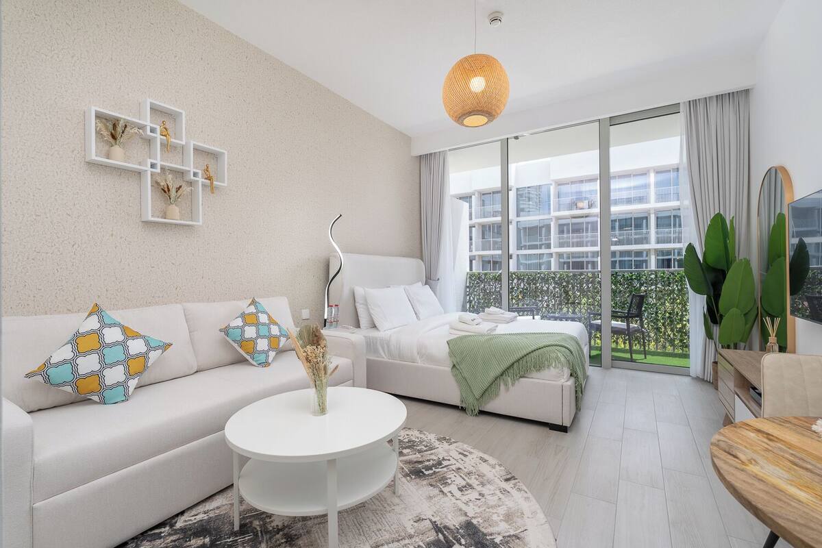 A modern living room with a white sofa, a round coffee table, and a bed with a green blanket. There is a large window with a view of buildings and a balcony.