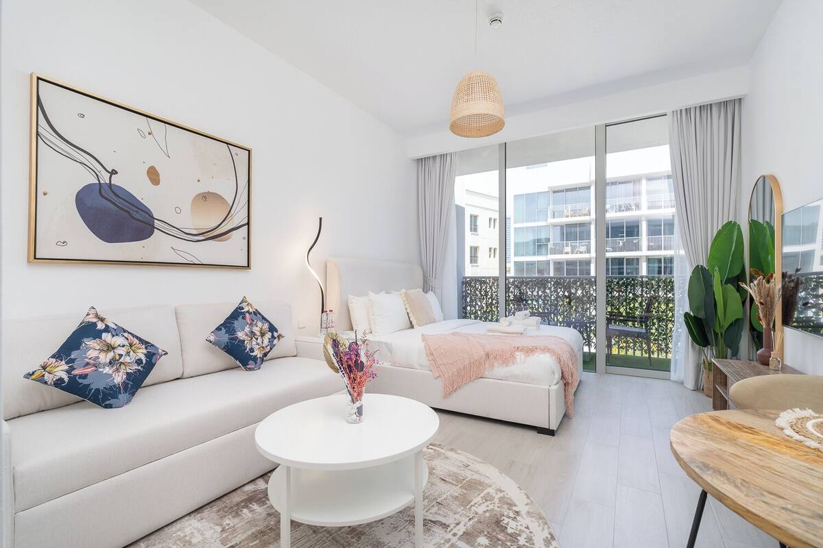 A modern bedroom with a large bed, a white sofa, a round coffee table, and a balcony with a view of the city.
